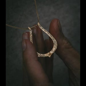 Puede incluir: Un collar con colgante en tono dorado que presenta un marco en forma de lágrima con un patrón de greca repetitivo. El colgante está suspendido de una delicada cadena dorada, mostrando un acabado metálico pulido y diseño geométrico.