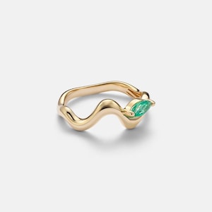 May include: A gold ring featuring a smooth, undulating wave-like band. One end of the band is set with a single, small, marquise-cut green gemstone, which is positioned to rest along the finger when worn. The metal has a polished, reflective finish.