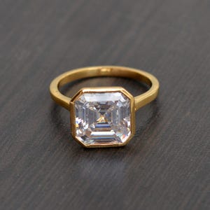Puede incluir: Un anillo de oro con una gran piedra preciosa transparente de talla cuadrada. La piedra preciosa está engastada en un bisel y la banda es de oro pulido simple. El anillo se muestra sobre un fondo oscuro.