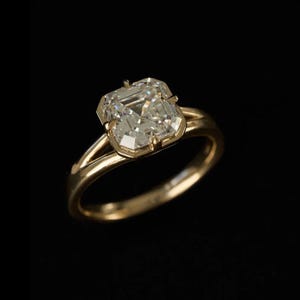 May include: A gold ring featuring a square-cut, faceted clear gemstone set in a four-prong mounting. The band has a split-shank design where the metal separates as it reaches the central stone, creating a polished and reflective metallic finish.