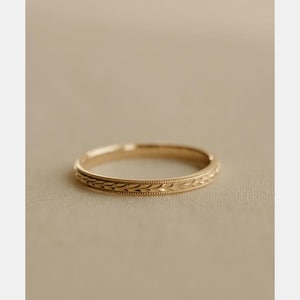 Op de afbeelding: Een dunne gouden ring met een doorlopend gegraveerd bladpatroon rond de buitenste omtrek. De ring is afgewerkt met delicate kralenachtige milgrain-details langs zowel de boven- als onderrand van de metalen band.