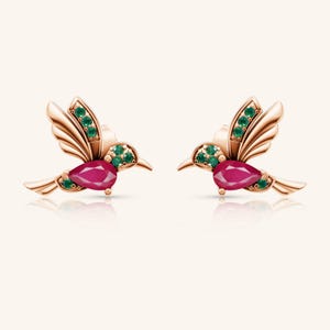 May include: A pair of hummingbird earrings. Each earring features a rose gold-toned metal hummingbird design with a pear-shaped ruby gemstone body and emerald green gemstone accents on the wings and head. The earrings are displayed on a light background.