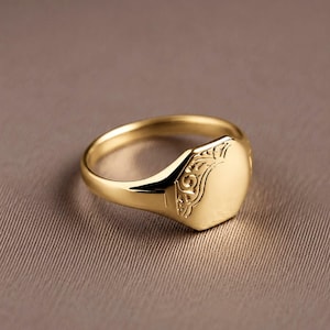 Op de afbeelding: Een gouden zegelring met een glad, plat, zeshoekig vlak en een gepolijste ringband. De zijkant van de ring is versierd met een ingewikkeld, gegraveerd krulpatroon dat zich uitstrekt van de ringband naar het centrale vlak van het sieraad.