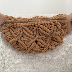 May include: A brown macrame fanny pack with a woven design. The bag has a zipper closure and a brown leather strap. The fanny pack is a neutral color and is suitable for everyday use.