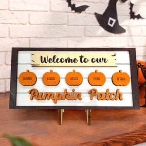 May include: A rectangular wooden sign with a dark frame and white shiplap background. It features the text "Welcome to our" and "Pumpkin Patch" in black and orange, with five orange pumpkin cutouts labeled with the names Damnis, Kalisei, Bessll, Fiona, and Jessica.