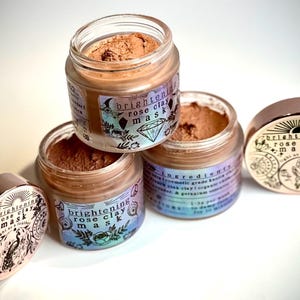 May include: Four small glass jars of rose clay mask with rose gold lids. The jars are filled with a light brown powder. The labels read "brightening rose clay mask" with a diamond and floral design.