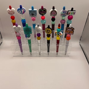 May include: A collection of decorative pens with various toppers. The toppers include heart shapes, rainbows, and text such as "Soul Sisters" and "Best Bonus Mom Ever." The pens are displayed in a clear acrylic stand.