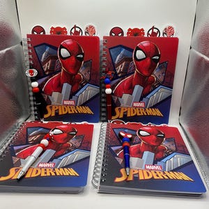 May include: Four spiral-bound notebooks featuring a red and blue Spider-Man graphic with the Marvel Spider-Man logo. Each notebook is paired with a matching pen topped with a Spider-Man character charm and colorful beads, displayed in a set.