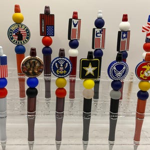 May include: A collection of pens with decorative tops featuring patriotic and military-themed designs. The pens have various colors, including red, white, blue, and gold, and include symbols like the American flag, stars, and military emblems.