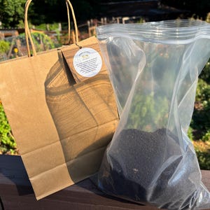 May include: A brown paper gift bag with a circular white tag featuring printed text and a clear plastic zip-top bag filled with dark black soil. The tag includes the text "Earthworm Castings" and additional descriptive information.