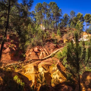 May include: Scenic landscape featuring a wooden walkway winding through a red and yellow rock formation. Tall green trees and a clear blue sky create a natural backdrop. The image captures a sunny day in a natural setting.