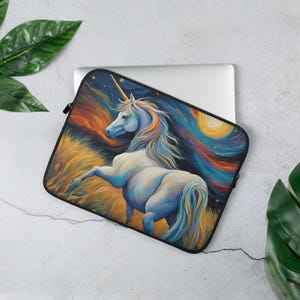 Peut inclure: Une housse pour ordinateur portable avec une peinture vibrante d'une licorne blanche avec une corne dorée, sur fond de ciel nocturne coloré et de champ d'herbes hautes. L'œuvre utilise des bleus, des jaunes et des oranges.
