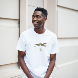 Puede incluir: Camiseta blanca con un diseño de ventilador de techo de dibujos animados. Las aspas del ventilador son marrones, el sol es amarillo y una bombilla cuelga debajo. La camiseta la lleva una persona.