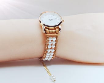 May include: A gold-toned wristwatch with a white face and pearl-studded band. The watch features a gold chain with pearls at the end. The watch is on a wrist.
