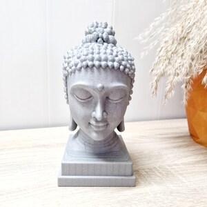 Puede incluir: Una estatua de cabeza de Buda gris con los ojos cerrados y una expresión serena. La estatua, con cabello detallado, se asienta sobre una base cuadrada. Se coloca sobre una superficie de madera clara. El fondo incluye una pared blanca y un jarrón decorativo.
