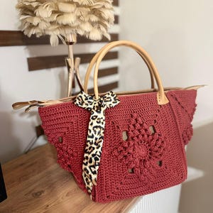 May include: A red crochet tote bag with a floral design and leather handles. The bag features a leopard print scarf tied to the handle. The bag is sitting on a wooden surface.