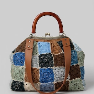 May include: A handcrafted crochet handbag with a wooden handle and brown leather straps. The bag features a patchwork design in shades of blue, brown, green, and black. The frame is a bronze color.
