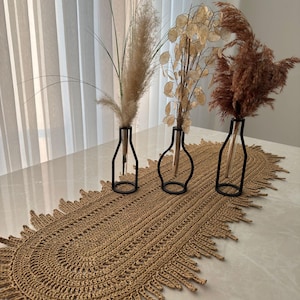 Könnte beinhalten: Eine Tischdekoration mit einem gewebten Tischläufer aus Jute mit Fransen. Drei Glasvasen mit schwarzen Metallständern halten getrocknete Ziergräser und Samenkapseln. Die Anordnung befindet sich auf einer hellen Oberfläche.