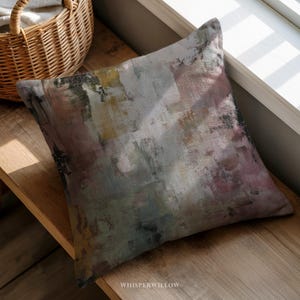 May include: Square decorative pillow with an abstract design in shades of gray, pink, yellow, and black. The pillow has a textured appearance and is placed on a wooden surface near a window. The text "WHISPERWILLOW" is visible.
