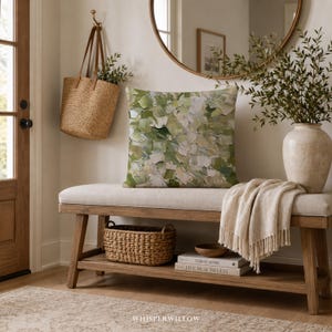 May include: A square throw pillow featuring an abstract impasto-style oil painting pattern in shades of sage green, olive, cream, and white. The pillow is placed on a light-colored upholstered wooden bench with a woven basket and books below.