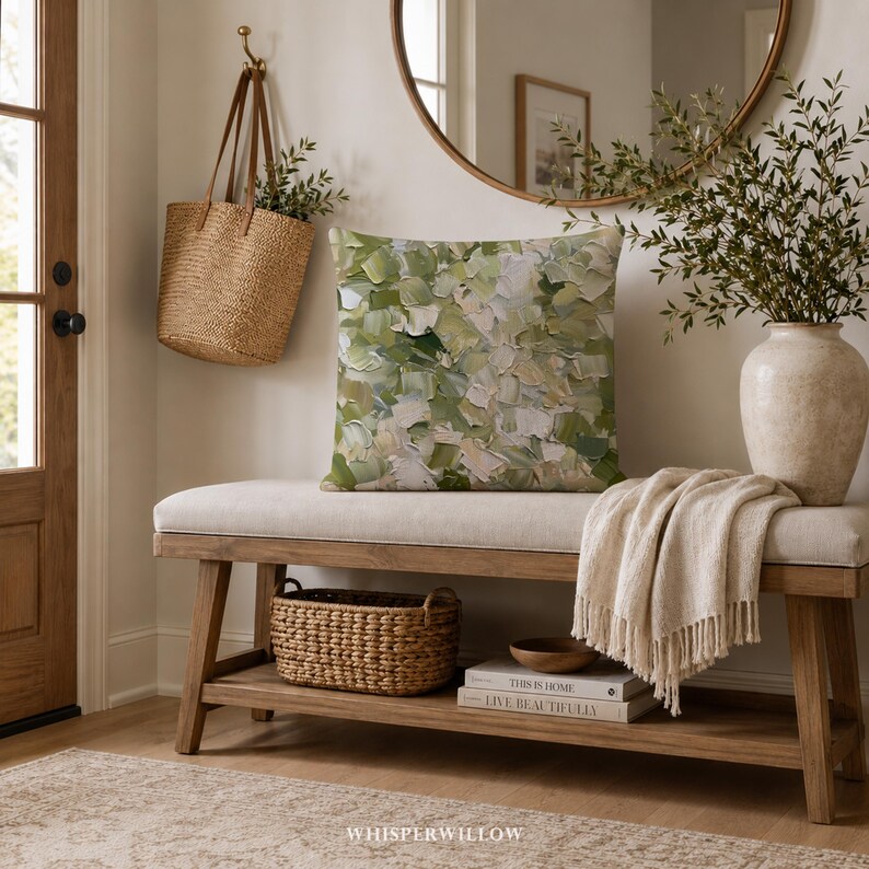 May include: A square throw pillow featuring an abstract impasto-style oil painting pattern in shades of sage green, olive, cream, and white. The pillow is placed on a light-colored upholstered wooden bench with a woven basket and books below.
