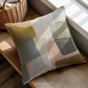 May include: Square decorative pillow with a geometric pattern in shades of green, gray, pink, and gold. The pillow is placed on a wooden surface near a window. The text "WHISPERWILLOW" is visible.