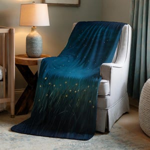 Puede incluir: Una manta azul oscuro sobre un sillón blanco. La manta presenta un diseño de hierba alta y orbes amarillos brillantes, con un patrón de cielo estrellado. La manta está hecha de material suave.