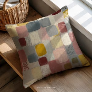 May include: Square throw pillow with a colorful abstract pattern. The design features overlapping squares in shades of yellow, pink, blue, and red. The pillow is resting on a wooden surface near a window.