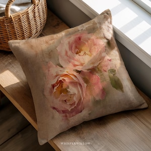 May include: Square decorative pillow with a floral design. The pillow features a watercolor painting of pink roses with yellow centers on a beige background. The pillow is made of a textured fabric and is placed on a wooden surface.