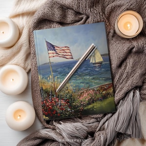 May include: A journal with an American flag painting on the cover, a silver pen, and three lit candles. The journal's cover depicts a coastal scene with a sailboat. The scene is set on a knitted blanket.