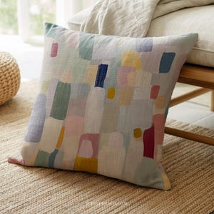 Puede incluir: Cojín decorativo cuadrado con un diseño abstracto colorido. El cojín presenta un patrón de formas rectangulares en varios colores pastel, incluyendo azul, rosa, amarillo y verde, sobre un fondo claro. El cojín está colocado sobre una alfombra tejida.