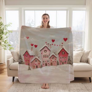 May include: A soft blanket with a winter village scene, featuring pink and red houses, snow, and red heart motifs. The blanket is displayed in front of a window and a sofa.