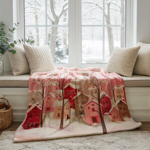 May include: A pink and white blanket featuring a winter village scene with red and pink houses, trees, and snow. The blanket is draped over a window seat with pillows. The text "WHISPERWILLOW" is visible on the blanket.