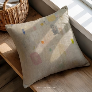 May include: Square decorative pillow with a light beige linen cover. The pillow features colorful abstract shapes in yellow, blue, pink, orange, and green. The pillow is resting on a wooden surface near a window.