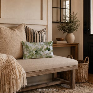 May include: A rectangular throw pillow featuring an abstract painterly pattern in shades of sage green, olive, cream, and white. The pillow is positioned in front of a large, textured beige square pillow and a striped black and beige pillow.