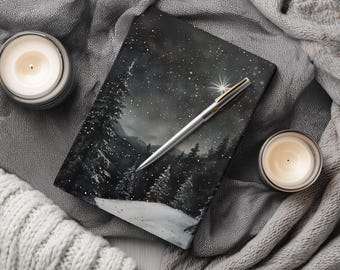 May include: A notebook with a winter landscape painting featuring a starry night sky, snow-covered ground, and evergreen trees. A silver pen with gold accents rests on the notebook. Two lit candles and a gray knitted scarf surround the notebook.