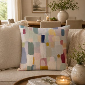 Peut inclure: Un coussin carré présentant un motif abstrait de coups de pinceau verticaux et rectangulaires arrondis dans des tons vert sauge, rose poudré, jaune moutarde, bleu marine et crème sur un fond en tissu texturé beige clair.