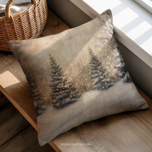 May include: Square decorative pillow with a winter forest scene. The pillow features a print of snow-covered evergreen trees with twinkling lights. The pillow has a neutral color palette with a linen-like texture. The text "WHISPERWILLOW" is printed on the bottom.
