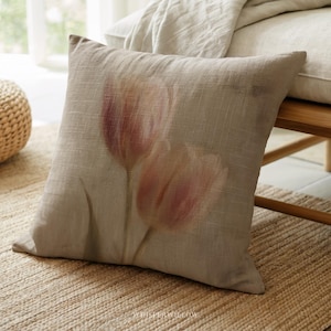 May include: A square beige pillow with a floral design. The pillow features a soft, muted image of two pink tulips. The pillow is made of a textured fabric and is placed on a woven rug.