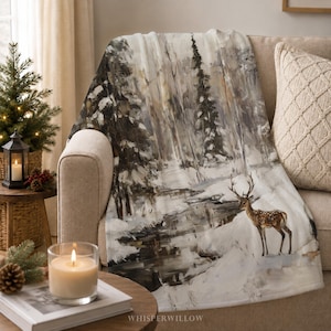 Könnte beinhalten: Eine weiche Wohndecke mit einer malerischen Winterlandschaft, die einen verschneiten Wald, einen dunklen Bachlauf und einen braunen Hirsch im Vordergrund zeigt. Das Motiv verwendet gedeckte Töne in Weiß, Grau, Braun und Schwarz.