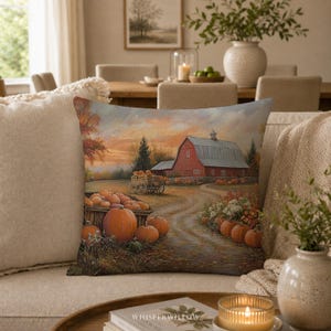 Op de afbeelding: Een vierkant sierkussen met een bedrukte landschapsafbeelding van een rode schuur, een houten kar vol oranje pompoenen en een zandpad. Het herfstachtige kunstwerk toont een zonsondergang, groene bomen en pompoenen op de grond.