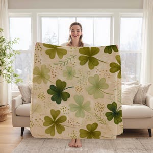 May include: A beige blanket with a pattern of green shamrocks and floral accents. The blanket is held up in front of a window, showcasing the design. The blanket is a cozy home decor item.