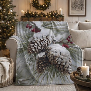 Könnte beinhalten: Eine weiche Wohndecke mit einem detaillierten Druck von zwei großen, braunen und schneebedeckten Tannenzapfen, eingebettet zwischen grünen Tannennadeln und kleinen roten Beeren auf einem hellen, winterlichen Hintergrund mit Schneetextur.