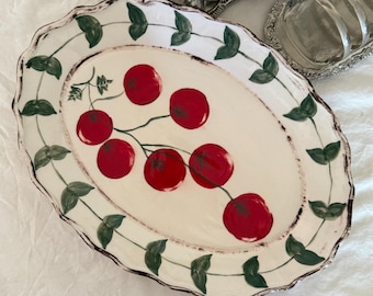 May include: Oval ceramic serving platter with a cream-colored center and a scalloped edge. The platter features a hand-painted design of red cherries on a branch, with green leaves and a brown rim.