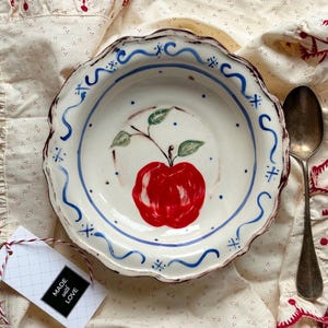 Op de afbeelding: Een witte keramische kom met een rode appelillustratie en blauwe accenten. De kom heeft een geschulpte rand en ligt op een crèmekleurige doek. Een lepel ligt naast de kom en een label met "Made with Love" is bevestigd.
