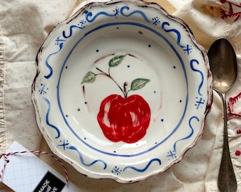 May include: A white ceramic bowl with a red apple illustration and blue accents. The bowl has a scalloped edge and is on a cream-colored cloth. A spoon rests beside the bowl, and a tag that says "Made with Love" is attached.