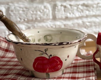 May include: Cream-colored ceramic bowl with a large red apple design, a handle, and a wooden spoon. The bowl sits on a red and white checkered cloth. The rim of the bowl has a distressed, rustic look.