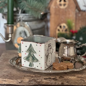 May include: A square ceramic Christmas-themed container with a green Christmas tree and a snowman design. The container is white with red and black polka dots. A silver tray holds the container, a small silver pitcher, and gingerbread cookies.