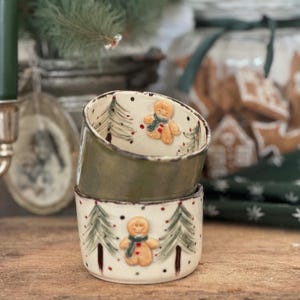 Könnte beinhalten: Zwei gestapelte Keramikschalen mit Weihnachtsmotiv. Die Schalen sind cremefarben mit grünen Akzenten und zeigen handgemalte Weihnachtsbäume, rote Punkte und Lebkuchenmänner mit Schals. Die obere Schale ist olivgrün.
