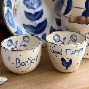 May include: Two small, off-white ceramic bowls with blue floral designs and brown rims. One bowl says "Bonjour" in blue script, the other says "Good Morning" with a blue rooster illustration. Other matching tableware is in the background.